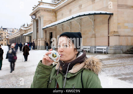 Junge Frau trinkt von Tasse mit therapeutischen Mineralwasser bei einer natürlichen heißen Quelle in Karlsbad im Winter, Tschechische Republik Stockfoto