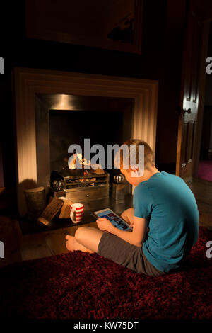 Teenager auf der Suche nach Tablet vor dem Feuer. Hygge Stockfoto