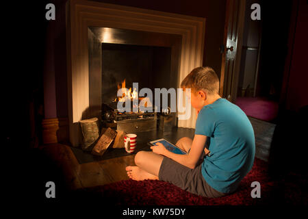 Teenager auf der Suche nach Tablet vor dem Feuer. Hygge Stockfoto