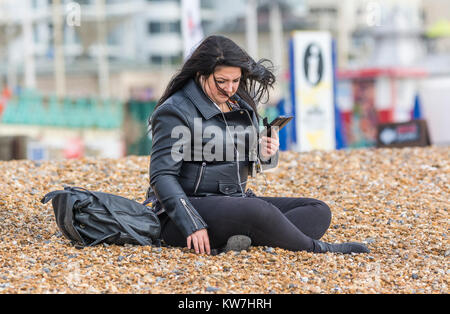 Junge Frau in Schwarz gekleidet sitzen allein auf einen Kiesstrand Hören von Musik über Kopfhörer, in Großbritannien. Stockfoto