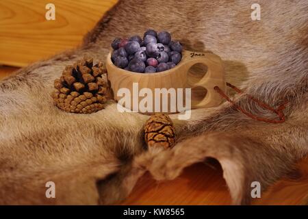 Heidelbeeren im Finnischen Holz- kuksy Schale auf ein Rentier Fell mit Kiefernzapfen. Nordeuropäischen noch Leben. Winter 2017. Stockfoto