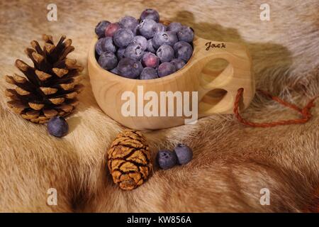 Heidelbeeren im Finnischen Holz- kuksy Schale auf ein Rentier Fell mit Kiefernzapfen. Nordeuropäischen noch Leben. Winter 2017. Stockfoto