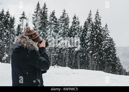 Junger Mann im Winter Mantel, Mütze und Schal, Foto mit DSLR-Kamera in Bukovel, Ukraine gekleidet, in einer kalten, schneereichen Winter morgen. Stockfoto