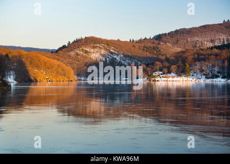 Abendlicher Blick über den zugefrorenen Obersee Rursee mit Reflexion auf die beleuchtete Landschaft im Winter in der Eifel. Stockfoto