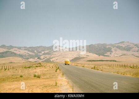 Ein gelber Bus fährt entlang einer Landstraße, die Kürzungen durch eine Wiese in trockenem Gras bedeckt, niedrigen Hügeln im Hintergrund, 1960 gesehen werden kann. Stockfoto