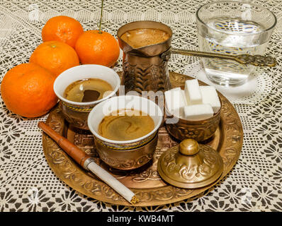 Türkischer Kaffee serviert mit einer Kaffeekanne, Würfelzucker und Glas Wasser mit Mandarinen und Zigarettenspitze auf weißen Deckchen Stockfoto