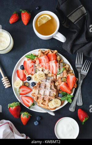 Hausgemachten belgischen Waffeln mit frischen Erdbeeren, Bananen und Heidelbeeren serviert mit Tasse grünen Tee mit Zitrone. Ansicht von oben, dunklen Hintergrund Stockfoto