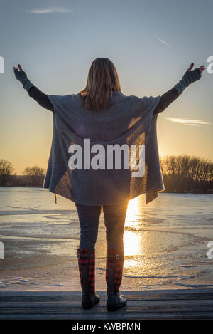 Betende Frau mit der Waffe in der Luft bei Sonnenuntergang im Winter auf dem zugefrorenen See Stockfoto