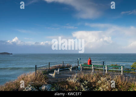 Eine einsame Frau, die Frau trägt eine rote Jacke, die auf einer Bank sitzen auf einer Aussichtsplattform mit Blick auf das Meer in Newquay Cornwall. Stockfoto