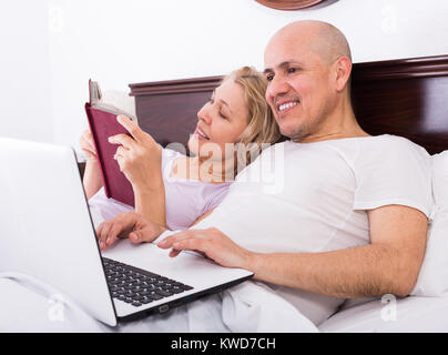 Freudige reifer Mann und Frau im Bett mit Laptop und Buch und lächelnd. Fokus auf den Menschen Stockfoto