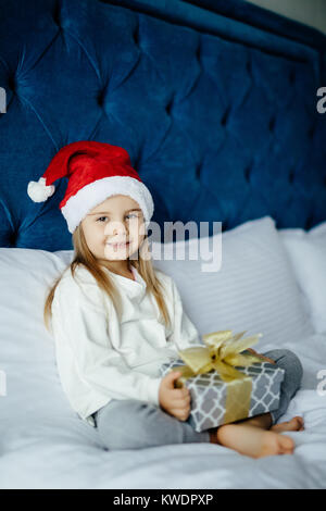 Happy kleines Mädchen in Santa's Hut holding Geschenkbox, an der Kamera schaut beim Sitzen auf dem Bett Stockfoto