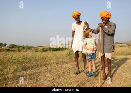 Western blonde Junge und zwei Kamel Händler mit orangefarbenen Turbanen und ihre Herden weiden im Busch in der Nähe des jährlichen Pushkar Camel Fair, Rajasthan, Indien Stockfoto