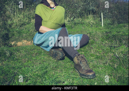 Eine junge Frau mit einem Baby im Tragetuch sitzt auf dem Gras außerhalb Stockfoto