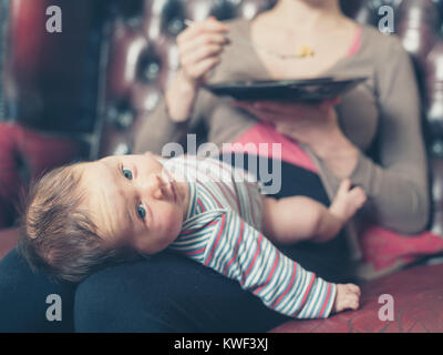 Eine junge Mutter sitzt auf einem Sofa und versucht ihr Abendessen mit Ihrem Baby auf dem Schoß zu essen Stockfoto