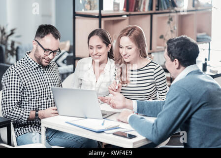 Lassen Sie mich erklären. Schöne stilvolle lächelnde Angestellte im Büro durch die Tabelle mit den Laptop und hören auf die Kollegen. Stockfoto