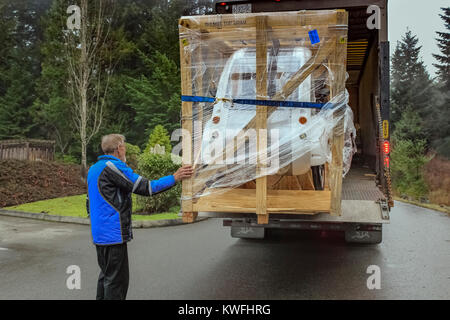 Ein älterer Mann in einen blauen Mantel Versand berührt eine große Kiste mit eine neue elektrische ELF Dreirad, als er für einen Transport LKW zu entladen wartet. Stockfoto