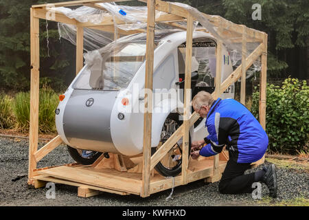 Ein älterer Mann in einem blauen Mantel kniet auf den Boden und nimmt mit einem Werkzeug eine hölzerne Transportkiste mit seinem neuen elf-Elektro-Dreirad auseinander. Stockfoto