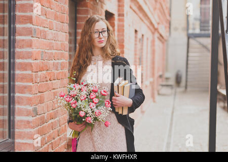 Ein curly - behaarte Frau mit Brille hält einen Strauß Tulpen und Paket der Bücher gegen den roten Ziegelgebäude Stockfoto
