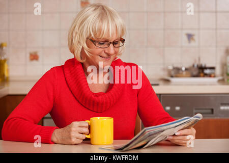 Ältere Frau, die einen Bruch in ihrer Küche das Lesen einer Zeitschrift und trinken eine Tasse Tee oder Kaffee. Stockfoto