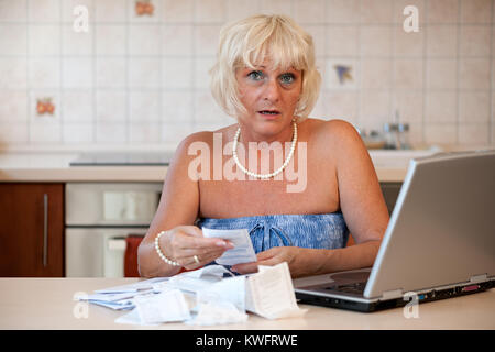 Schockiert Frau mittleren Alters in ihrer Küche am Tisch mit einem Laptop sitzen und die Kontrolle über die Preise auf die Einnahmen aus dem Supermarkt Stockfoto