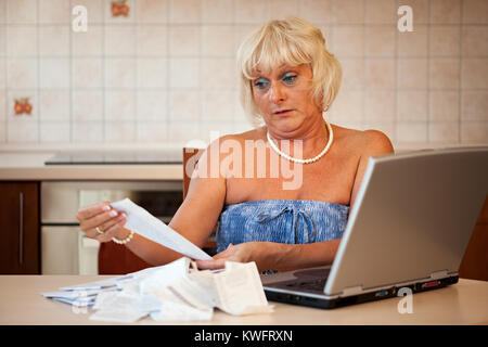 Schockiert Frau mittleren Alters in ihrer Küche am Tisch mit einem Laptop sitzen und die Kontrolle über die Preise auf die Einnahmen aus dem Supermarkt Stockfoto