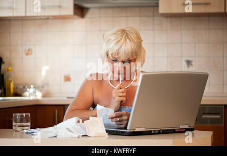 Schockiert Frau mittleren Alters in ihrer Küche am Tisch mit einem Laptop sitzen und die Kontrolle über die Preise auf die Einnahmen aus dem Supermarkt Stockfoto