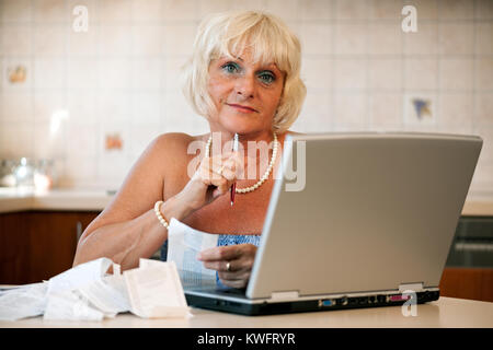 Gerne Frau mittleren Alters in ihrer Küche am Tisch mit einem Laptop sitzen und die Kontrolle über die Preise auf die Einnahmen aus dem Supermarkt Stockfoto