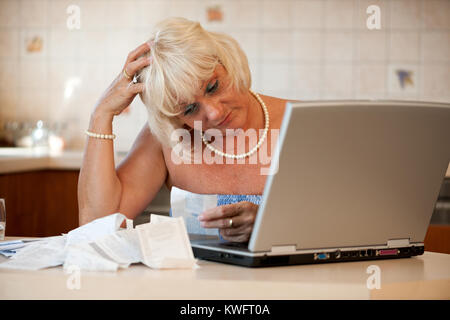 Schockiert Frau mittleren Alters in ihrer Küche am Tisch mit einem Laptop sitzen und die Kontrolle über die Preise auf die Einnahmen aus dem Supermarkt Stockfoto