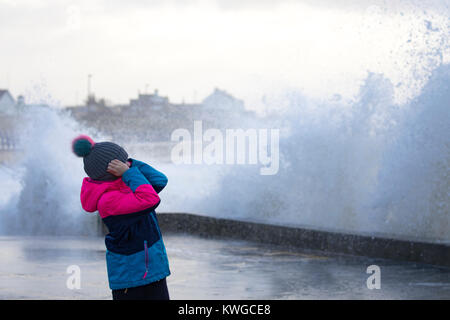 Anglesey, Wales, 3. Januar 2018. UK Wetter. Eine schwere Met Office Warnung hat für das fünfte Sturm der britischen Saison ausgestellt wurde Tropensturm Eleanor. Mit einem Anstieg der Gefahr durch die aktuelle Mond- und Gezeiten Hochwasserwarnungen für viele Bereiche zusammen mit gales Winde wahrscheinlich Schaden zu verursachen. Wellen und Wind Crash in Trearddur Bay auf Anglesey im Norden von Wales als junges Mädchen schützt Ihre Ohren von dem Lärm der Wellen an der Küste Stockfoto