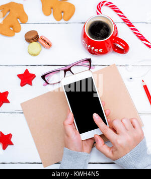 Zwei weibliche Hände halten ein Smartphone mit einem leeren Bildschirm schwarz, mit dem Finger auf den Bildschirm tippen, eine Sicht von oben, in der Nähe eines roten Tasse mit Kaffee Stockfoto