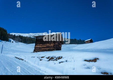 Alte Holzhütten und schneebedeckte Berge im Skigebiet Serfaus Fiss Ladis während der Nacht mit Mond und Sterne über Stockfoto