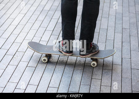 Eine Person in schwarzen Jeans und Trainer steht auf einem Skateboard auf einem gepflasterten Bereich Stockfoto