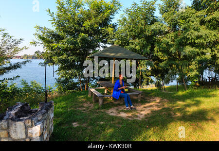 Glückliche Frau Tourist mit Compact Photo Kamera sitzt auf holzbank auf der malerischen Sommer Camping Stellplatz in der Nähe von See. Konzept der Umweltfreundliche Stockfoto