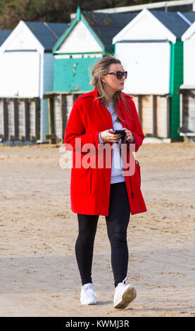 Bournemouth, Dorset, Großbritannien. Am 4. Januar, 2018. UK Wetter: windigen Tag am Bournemouth. Junge Frau im roten Mantel holding Handy Spaziergänge entlang der Promenade in Sand bei Alum Chine abgedeckt. Credit: Carolyn Jenkins/Alamy leben Nachrichten Stockfoto