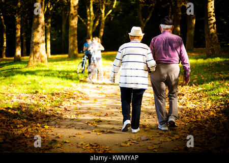 Ältere Paare gehen weg von der Kamera, Arm in Arm durch einen bewaldeten Park in einer Welle von Sonnenlicht unter den Bäumen Stockfoto