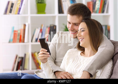 Glückliches Paar beobachten Medieninhalte auf einem Smartphone auf einer Couch zu Hause sitzen Stockfoto