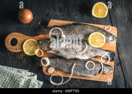 Oben Ansicht mit frischem Fisch gewürzt mit Gewürzen, Kräutern, Zitronenscheiben und Zwiebel Ringe auf eine hölzerne Schüssel, auf einem Schwarzen Tisch, in natürlichem Licht. Stockfoto