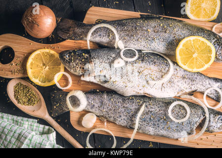 Close-up mit frischen Forellen, bestreut mit Kräutern, Gewürzen, Zwiebeln und Zitronenscheiben, auf einer hölzernen Schneidebrett platziert, auf einen Jahrgang Tabelle. Stockfoto