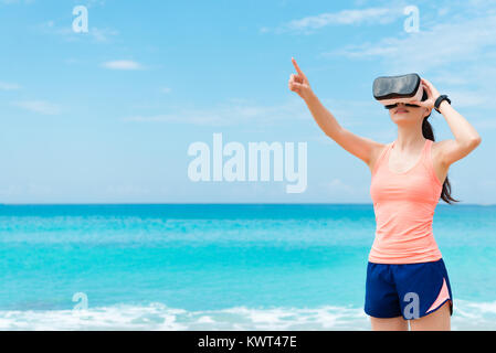 Schöne glückliche Frau Läufer tragen die VR-Technologie Brille, 3D-Video und zeigt mit der Hand berühren, Simulation, wenn sie Workout am Strand. Stockfoto