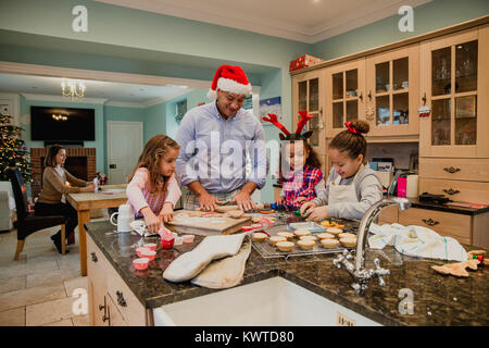 Kleine Mädchen machen Weihnachten Kekse zu Hause mit ihrem Vater. Die Mutter kann gesehen werden, Schreiben von Weihnachtskarten im Hintergrund. Stockfoto