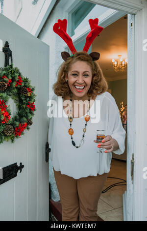 Sicht der Schuß eines fröhlichen älteren Frau heißen Sie in ihrem Haus für Weihnachten. Sie trägt Geweih und halten ein Glas Portwein. Stockfoto