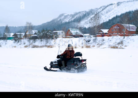 Ein Mann auf einem Schneemobil auf einem zugefrorenen Fluss vor dem Hintergrund einer verschwommenen Winter verschneite Landschaft im ländlichen Raum Stockfoto