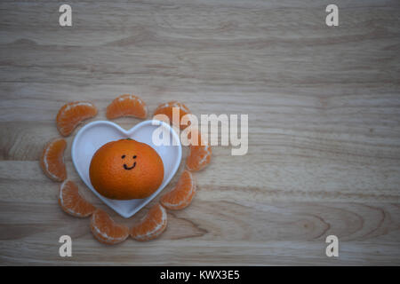 Essen Fotografie eines gesunden orange Satsuma oder Tangerine oder Clementine mit auf Lächeln und Obst Segmente mit einem weißen Liebe Herzform Teller Stockfoto