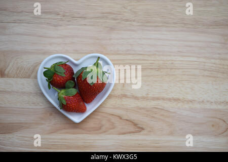 Essen Fotografie Bild von frischem Obst mit drei roten Erdbeeren mit grünem Blatt oben in einem weißen Liebe Herzform Teller auf Holz Hintergrund platziert Stockfoto