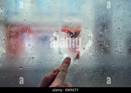 Finger des jungen Mannes Zeichnung Herz Form auf das Fenster mit Regentropfen gegen die Straße der Stadt. Stockfoto