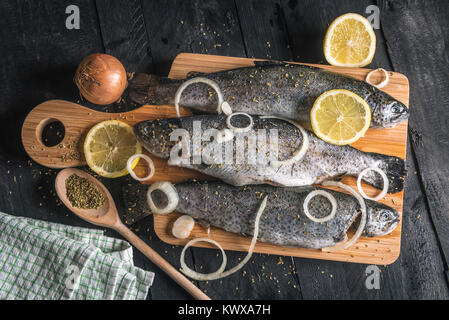 Hohe Betrachtungswinkel mit roher Fisch auf einem hölzernen Schneidebrett, gewürzt mit getrockneten Kräutern, Zitronenscheiben und Zwiebel Ringe, auf einem Vintage schwarz Tisch. Stockfoto