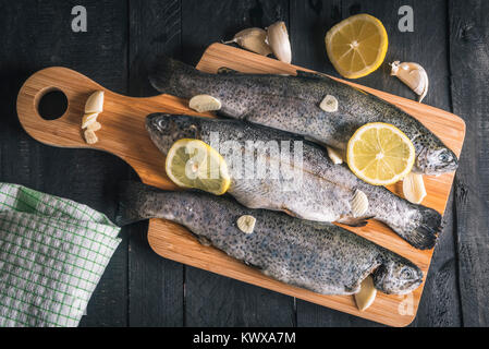 Oben Ansicht mit frischem Fisch mit Knoblauch gewürzt und Scheiben von Zitronen, auf einer hölzernen Schneidebrett angezeigt, auf einem urigen schwarz Tisch. Stockfoto