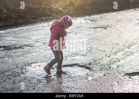 Konzept - glückliche Kindheit. Kleines Mädchen spielt in der Pfütze, Spaß für Kinder, schmutzige und nasse Schuhe, Leben im Dorf, Sonnenlicht, Ukraine Stockfoto
