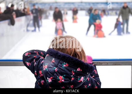 Ein junges Mädchen beugte sich über die Barriere an eine Eislaufbahn, beobachten die anderen Familien auf dem Eis mit skating Aids für die Kinder. Stockfoto