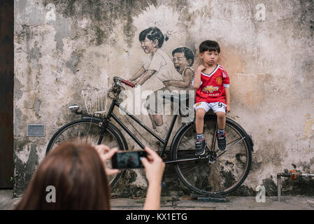 Eine Mutter nimmt ein Foto von ihrem Sohn auf einem Stück interaktive Street Art von Ernest Zacharevic in Georgetown, Penang, Malaysia sitzen. Stockfoto